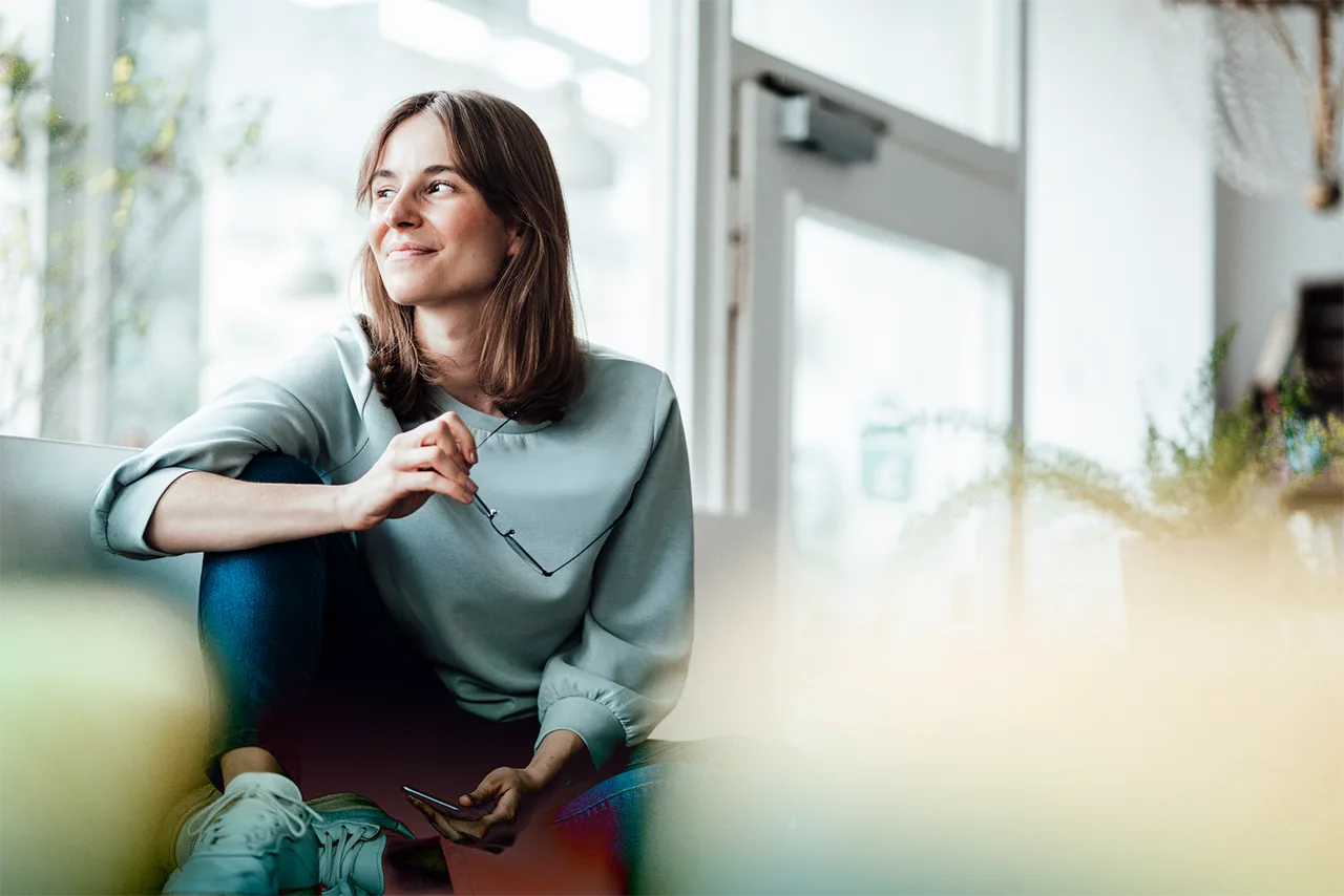 A woman sitting on a couch smiling and looking into the distance, holding a pair of glasses.