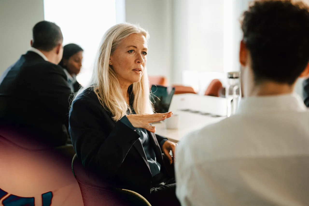 A blonde woman talking to a man at an office table, with colleagues in the background.